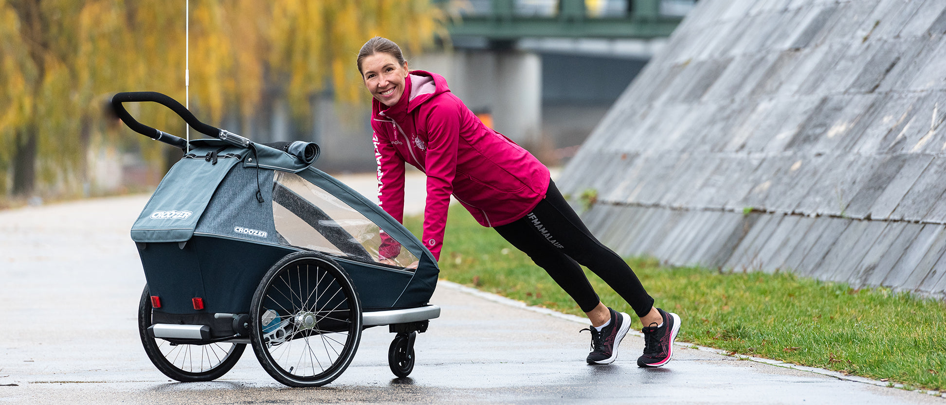 Smiling woman in incline push-up position against Croozer bike trailer with child inside during autumn workout